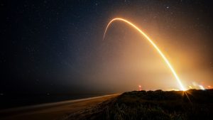 a time-lapse photo showing a streak from a rocket launch arcing over the beach at night.