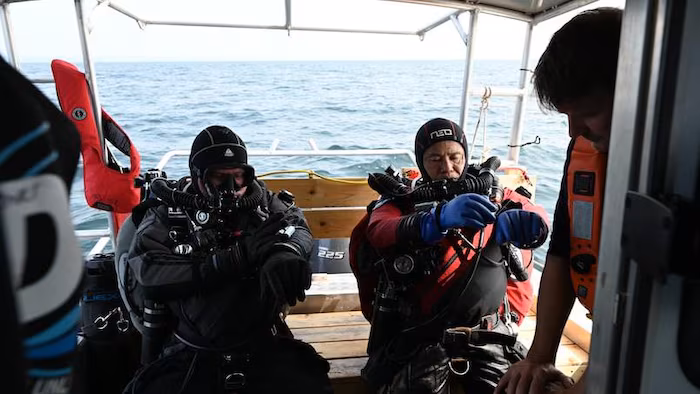 Trois hommes, dont deux portent de l'équipement de plongée, sur un petit bateau au milieu du lac.