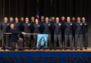 A group of people wearing dark blue shirts and black pants stand on stage with an American flag behind them and an officer kneeling down with a blue flag for the US Space Force placed in front of them.