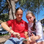 Omnia et Madeleine, assise sous un arbre. Omnia tient un portable, et fait un pouce en bas pour dire qu'elle n'approuve pas cette loi, et Madeleine fait un pouce en haut pour dire qu'elle, elle l'approuve.