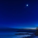 The moon is pictured glowing above the star-like form of Jupiter in a dark blue night sky above a snowy field bordering a road.