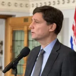 Un homme parle au micro. Derrière lui, le drapeau de la Colombie-Britannique.