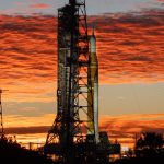 An orange rocket with white side booster stands next to the launch tower during a colorful sunrise.