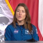 A woman with long dark hair wears a blue flight suit with the circular NASA logo on it and sits behind a desk with a microphone