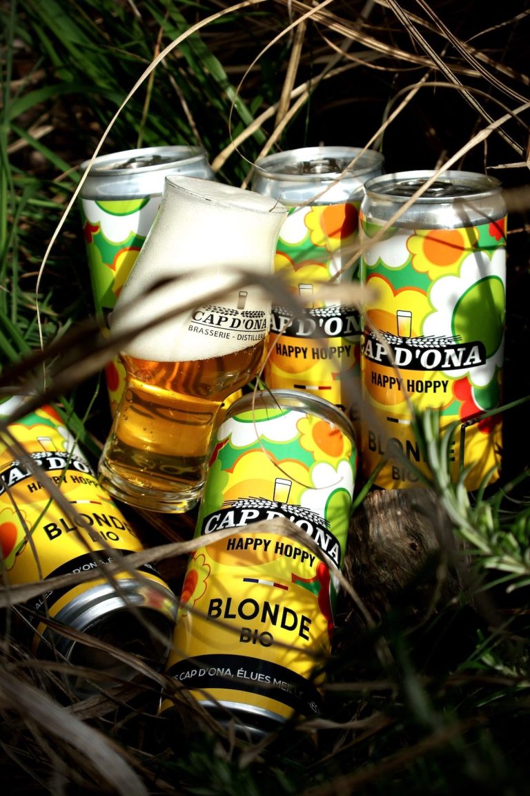 Des canettes de Happy Hoppy® Blonde Bio de Cap d’Ona entourent un verre de bière, image du nouveau format lancé par la brasserie de Céret.