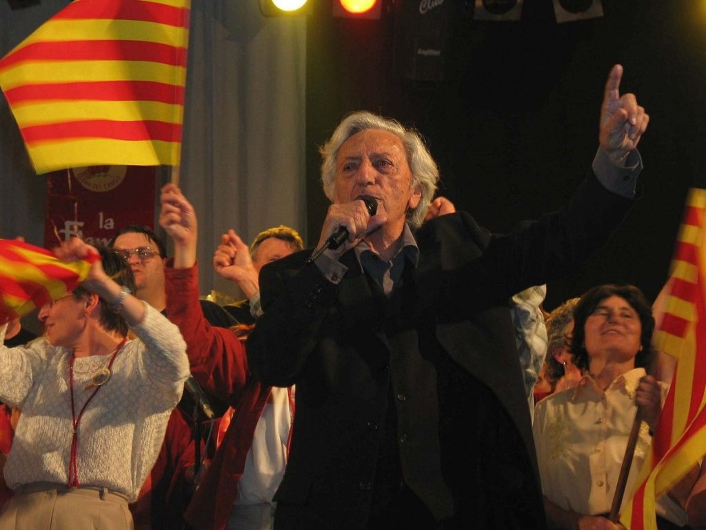 Jordi Barre chante au micro sur scène, entouré de plusieurs personnes brandissant des drapeaux catalans pendant une représentation publique.