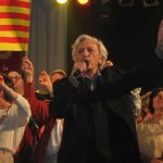 Jordi Barre chante au micro sur scène, entouré de plusieurs personnes brandissant des drapeaux catalans pendant une représentation publique.