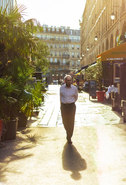 An adult man walks down a sunlit Parisian street, surrounded by urban architecture and greenery.
