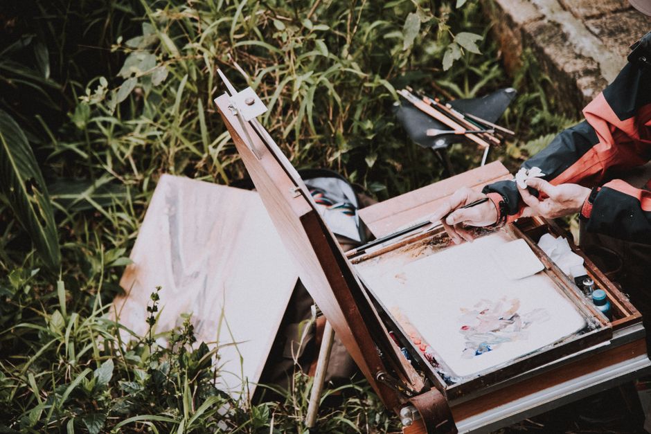 An artist painting outdoors with a wooden easel and canvas amidst lush greenery.