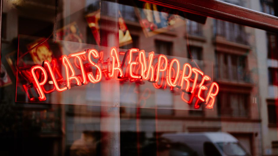 Red neon sign 'Plats à Emporter' reflected in city window, urban street vibe.
