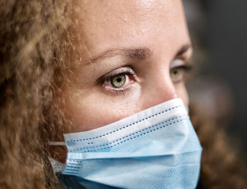 A woman with curly hair wears a medical face mask, highlighting her thoughtful eyes.