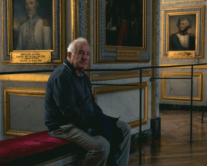 Elderly man seated on a bench in a Versailles art gallery, surrounded by historical paintings.
