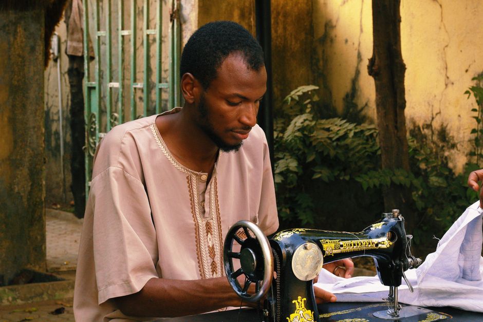 African male tailor focused on sewing with traditional machine in outdoor setting, Abuja, Nigeria.
