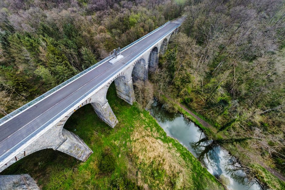 A drone captures a bridge over lush greenery and a serene river in Chimay, Belgium.