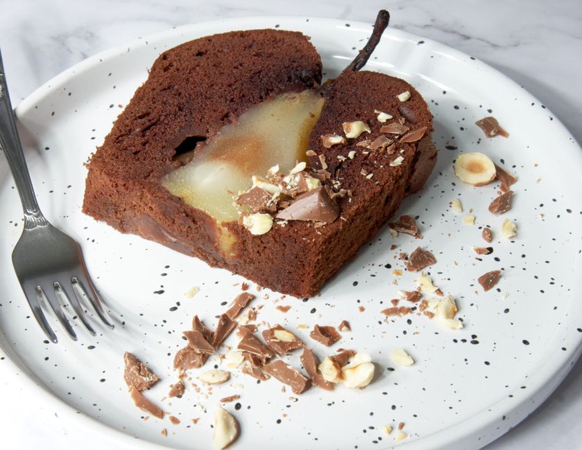 A slice of chocolate pear cake with hazelnuts on a speckled plate.