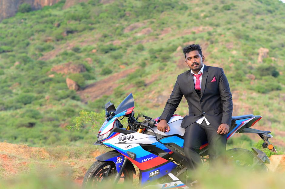 A man wearing a black suit stands by a Yamaha YZF-R15 V3 motorcycle in a scenic rural setting, ideal for adventure branding.
