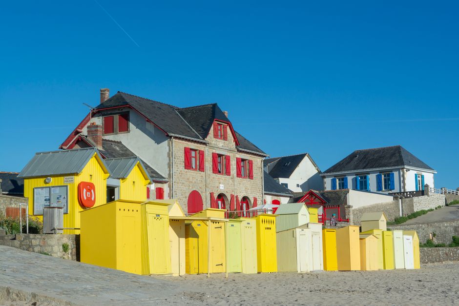 Vibrant beach huts line the sandy shore in Batz-sur-Mer, France, under a clear blue sky.