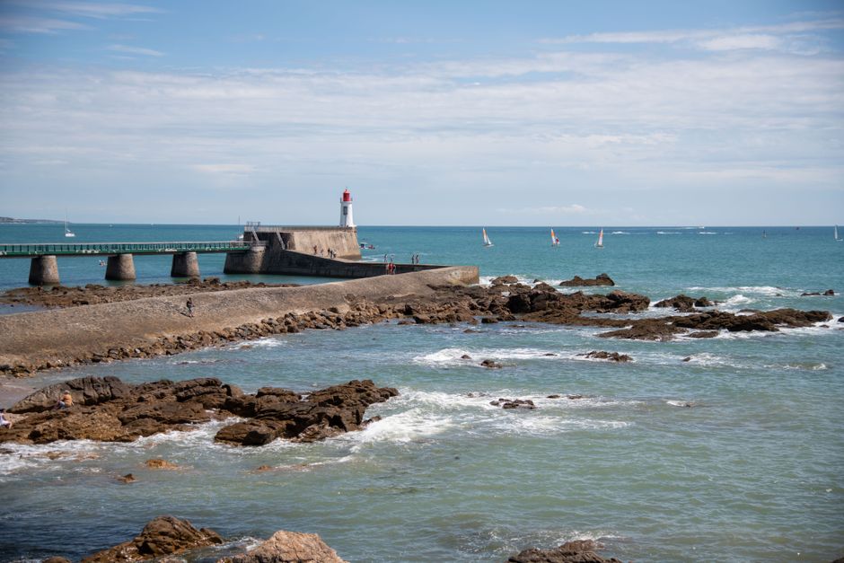 Picturesque view of a lighthouse by the sea with a rocky shore and sailboats on a sunny day.