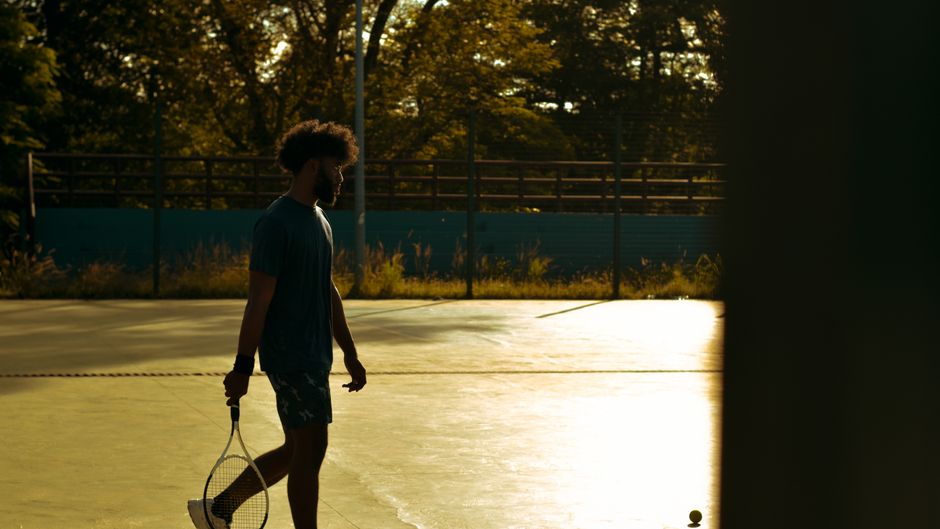 Man walks on a sunlit tennis court at sunset, holding a racket. Perfect lighting for sports imagery.