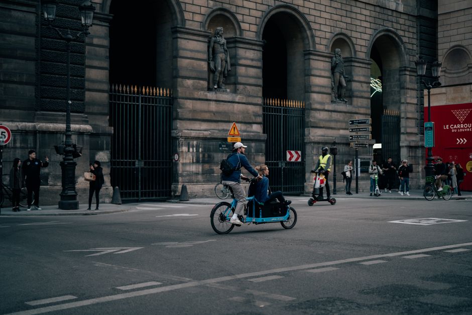A cyclist with a passenger and a scooter rider in front of the Le Carrousel du Louvre, Paris.