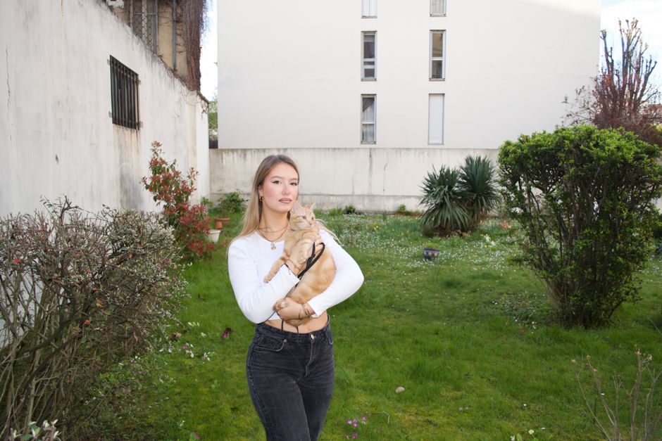 A woman stands in an urban garden holding a ginger cat on a leash, surrounded by greenery.
