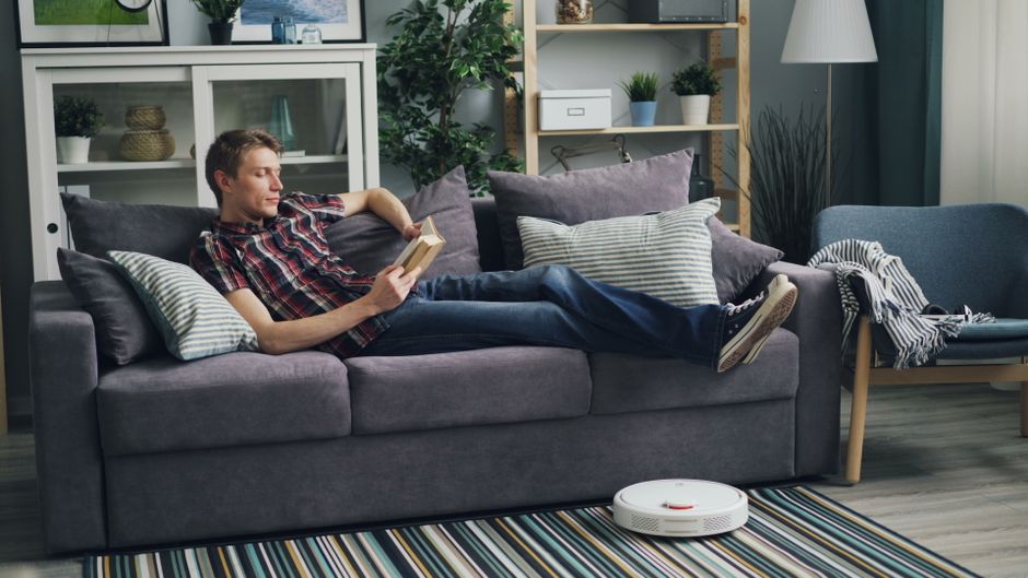 A man reading a book on a sofa, while a robotic vacuum cleans the living room floor.