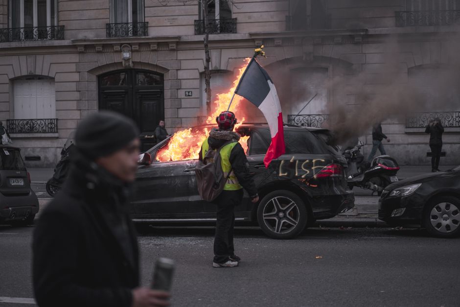 Man in yellow vest holding French flag near burning car during protest in Paris.