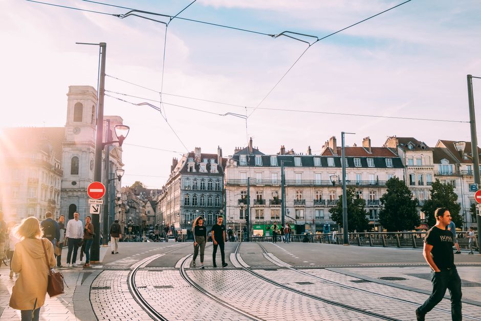 People walking across a sunny tramway intersection in a historic European city.
