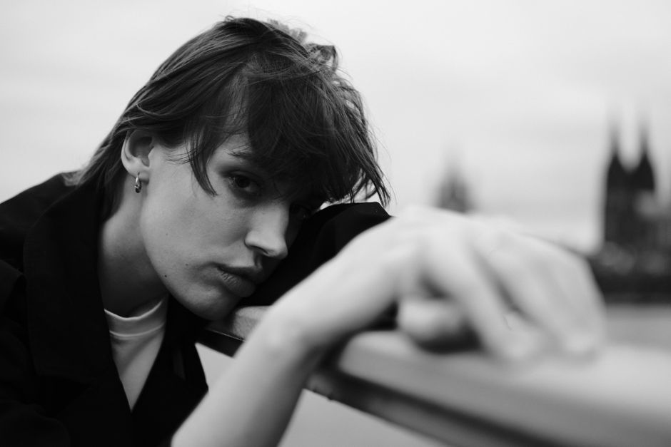 Black and white atmospheric portrait of a pensive adult leaning on a bridge railing in an urban setting.