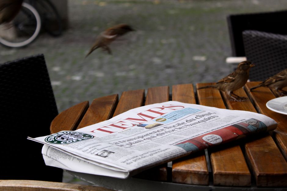A newspaper on a cafe table with sparrows nearby, creating a lively street scene.