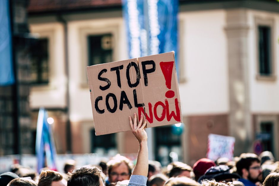 A crowd gathers for an outdoor protest with a sign reading 'Stop Coal Now'.