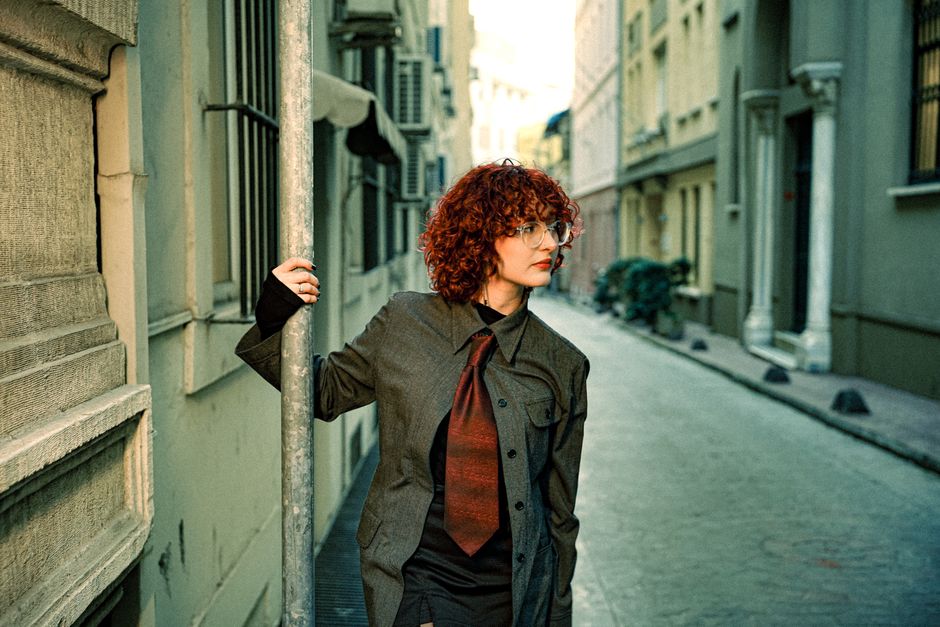 A fashionable woman with curly hair stands outdoors, holding a pole in a city alleyway.