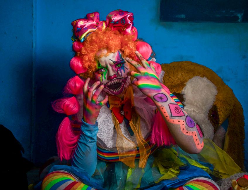 A person in a colorful clown costume with vibrant makeup posing indoors.