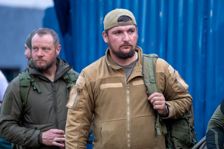 Two men wearing tactical gear standing outdoors against a blue background.