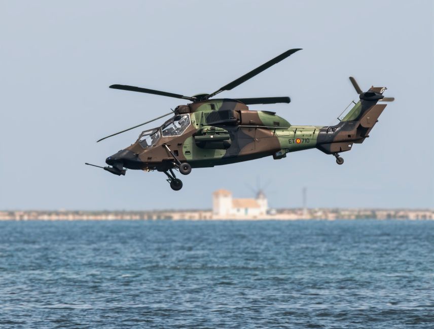 A camouflaged military helicopter hovering above the water with coastline visible in the background.