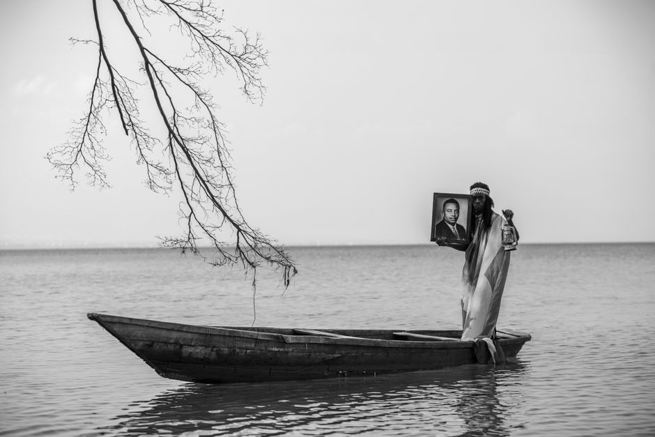 A black and white image of a person on a boat holding a portrait and lantern, with a dramatic branch overhang.