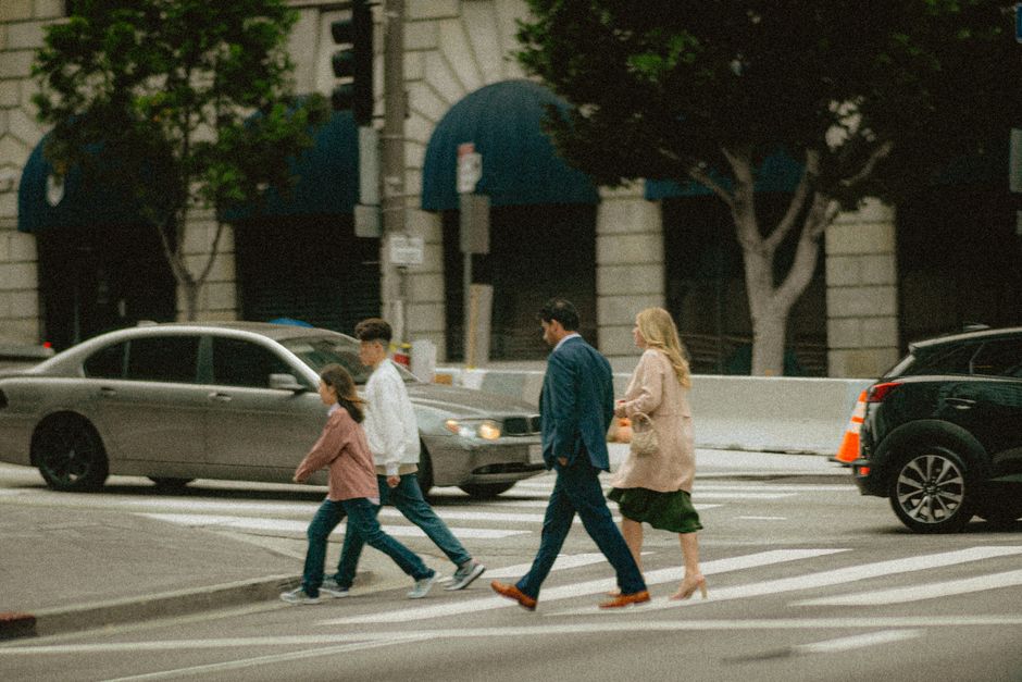 A family crosses a city street at a crosswalk surrounded by cars and buildings.