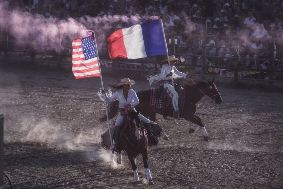 Dynamic rodeo scene with riders holding US and French flags under dusty arena.