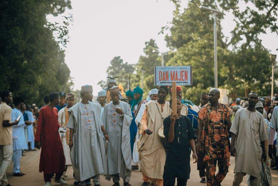 A vibrant cultural procession with a diverse group holding a 'Dan Majen' sign outdoors.