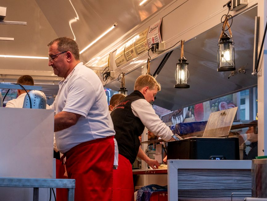 Street food vendor in red apron prepares meals inside a food truck. Busy evening scene.
