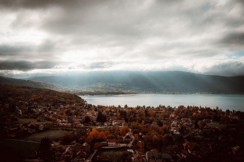 Breathtaking aerial view of Lake Annecy with autumn foliage and dramatic clouds.