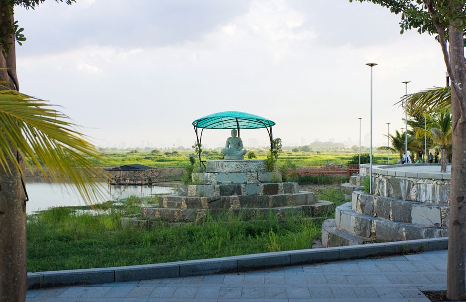 Peaceful riverside scene with a Buddha statue under a green shelter, surrounded by nature.