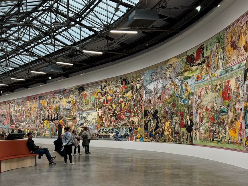 A large, colorful artwork displayed in a contemporary indoor gallery with people viewing.