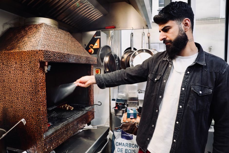 Bearded chef grilling meat skewers in a traditional Paris kitchen setting.