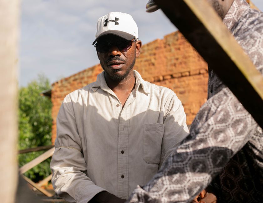 A man wearing sunglasses and a white cap stands outdoors in warm light.