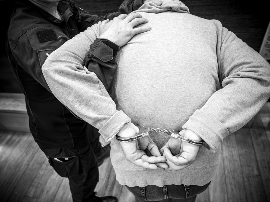 Black and white photo of a person being arrested indoors, handcuffed by an officer.