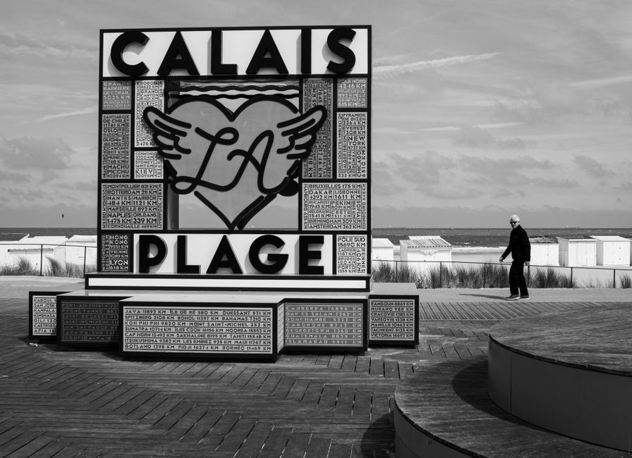 Black and white photo of Calais La Plage sign with cloudy skies and a lone person walking.