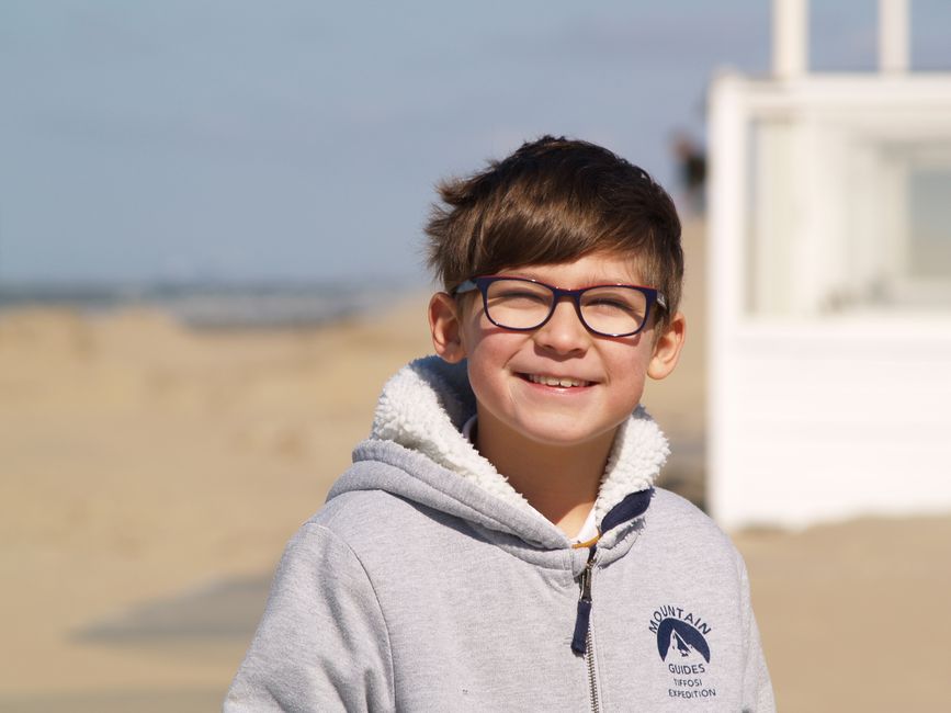 A cheerful child with glasses smiles at the beach, wearing a cozy hoodie on a sunny day.