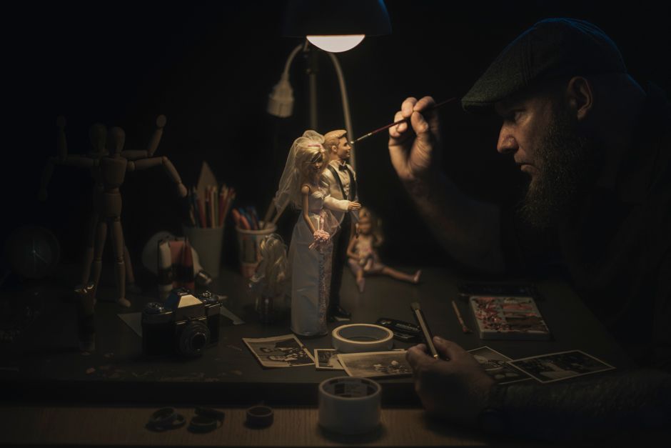 A craftsman meticulously painting bride and groom figurines under a lamp, focused on details.