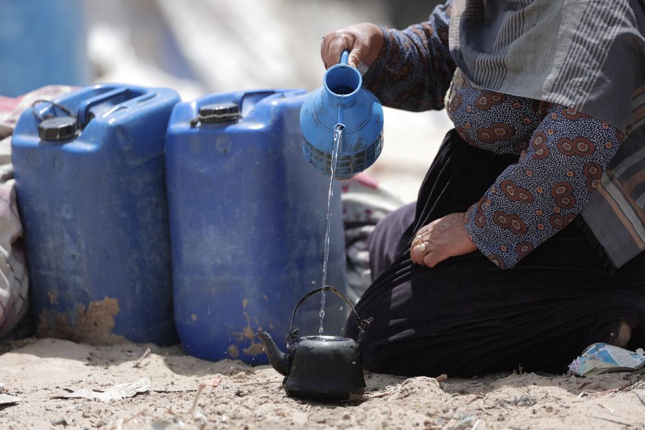 A woman pouring water into a kettle beside blue jerry cans, likely in the Middle East.